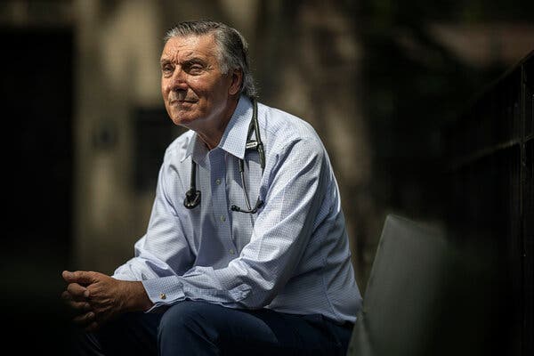 A man sits, in a button-down shirt with a stethoscope around his neck and his hands clasped in his lap.