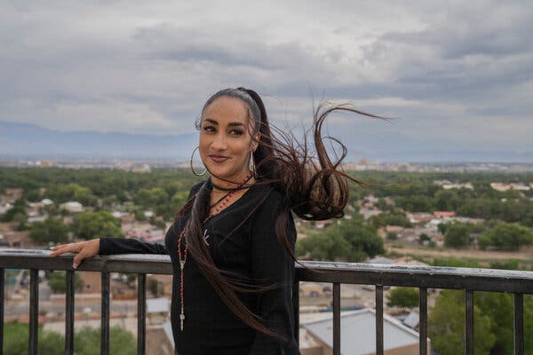 A woman stands outside, slightly smiling, with one hand resting on a railing and her long ponytail blowing in the wind.